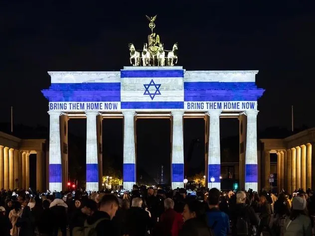 Studie Deutsch-Israelische Kulturbeziehungen Das Bild zur Studie Deutsch-Israelischer Kulturbeziehungen zeigt das Brandenburger Tor bei Nacht mit Projektion der israelischen Flagge und dem Text „BRING THEM HOME NOW“, umgeben von einer Menschenmenge.