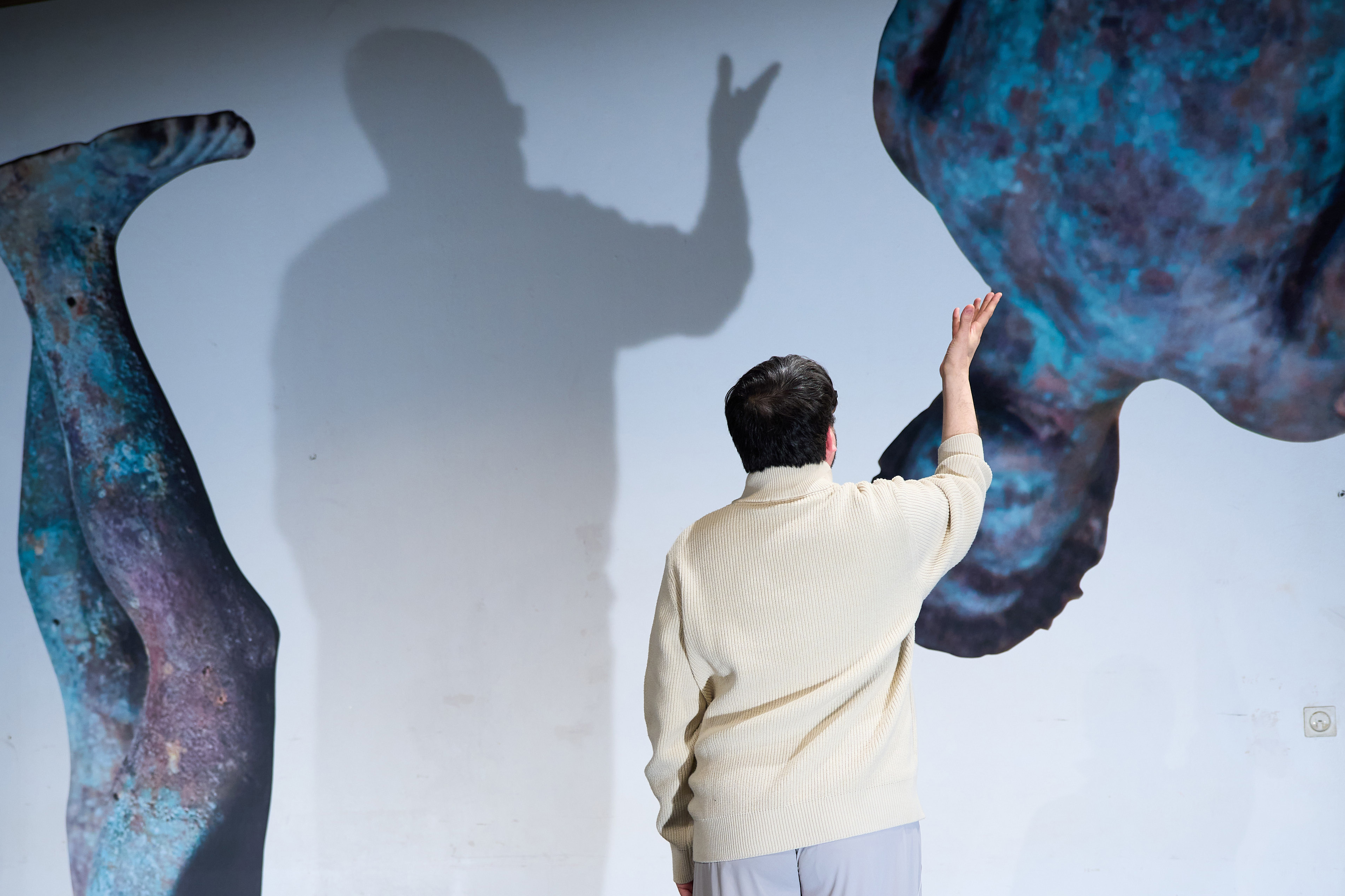Das Bild wurde aufgenommen während der Premiere des Theaterstücks Dualidarität. Es zeigt einen der beiden Schauspieler:innen, welcher vor einer Wand steht auf der Abbildungen von zwei großen, fragmentierten Apolloskulpturen zu sehen sind. Der Schauspieler wirft einen Schatten an die Wand, der eine Hand mit ausgestreckten Fingern zeigt.