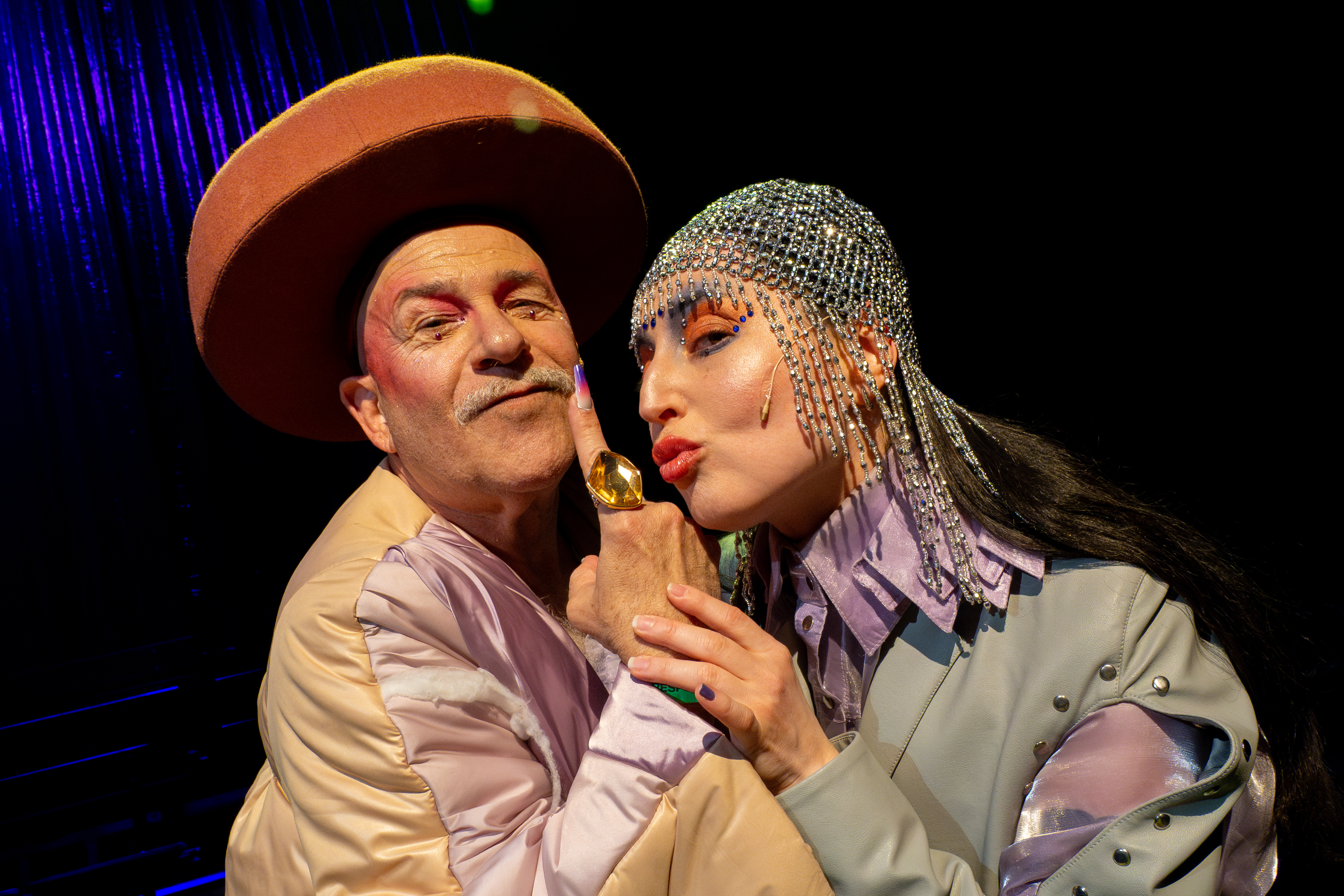 Das Bild wurde während des Purimshpils 2025 an den Münchner Kammerspielen aufgenommen und präsentiert die Performance-Serie House of Diaspora. Man sieht zwei Personen in auffälligen Kostümen, eine mit großem Hut und die andere mit glitzerndem Kopfschmuck. Bei den Personen handelt es sich um den König Achaschwerosch und Königen Esther.