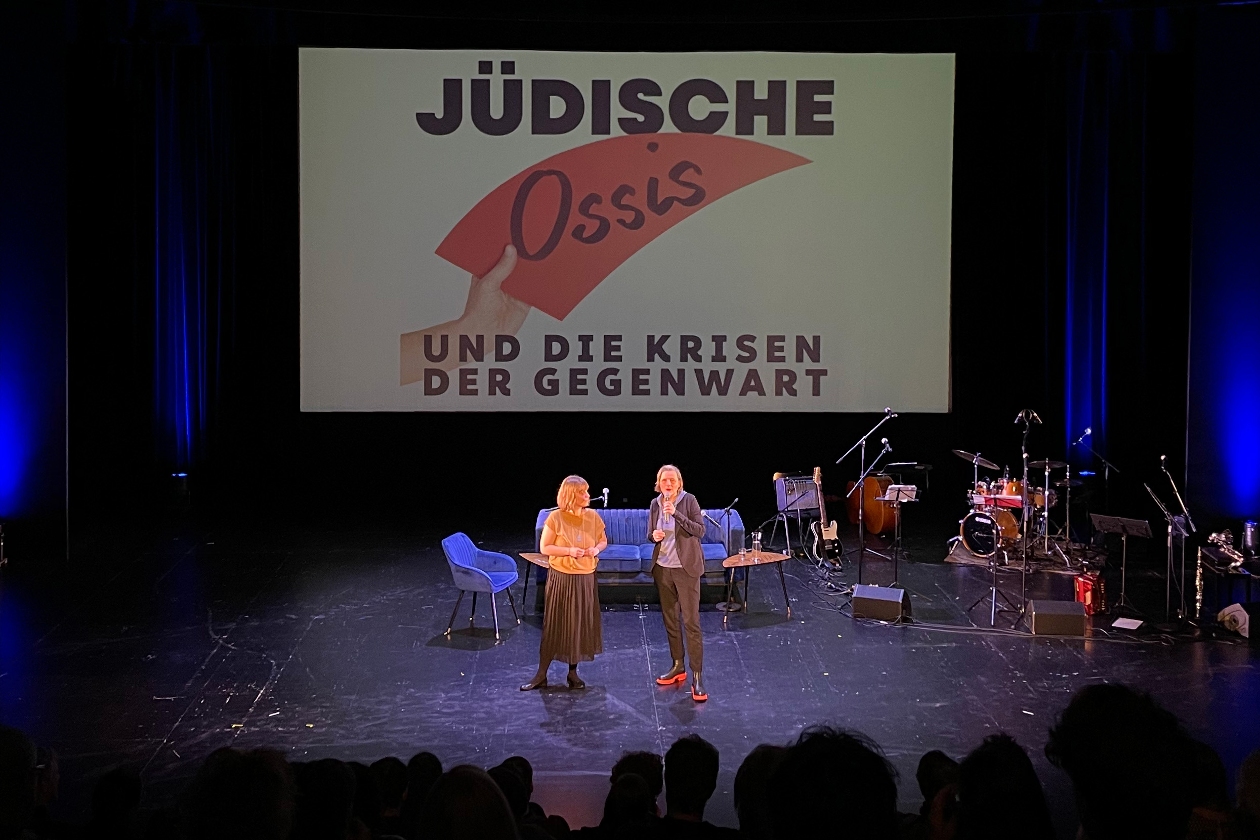 Zwei Personen stehen während der Veranstaltung »Jüdische Ossis« auf der Bühne des Hans Otto Theaters. Man sieht eine Leinwand mit dem Text »Jüdische Ossis und die Krisen der Gegenwart«, umgeben von Musikinstrumenten und Mikrofonen.