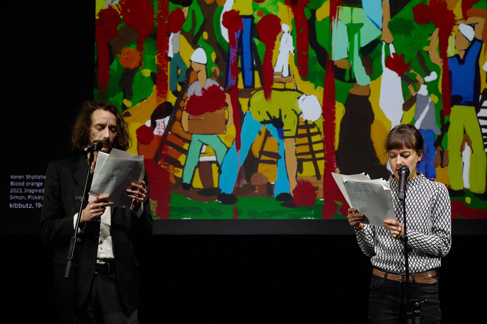 Das Bild zeigt zwei Personen die vor einer großen Leinwand stehen und in Mikrophone sprechen. Auf der Leinwand ist ein farbenfrohes, expressives Gemälde zu sehen.