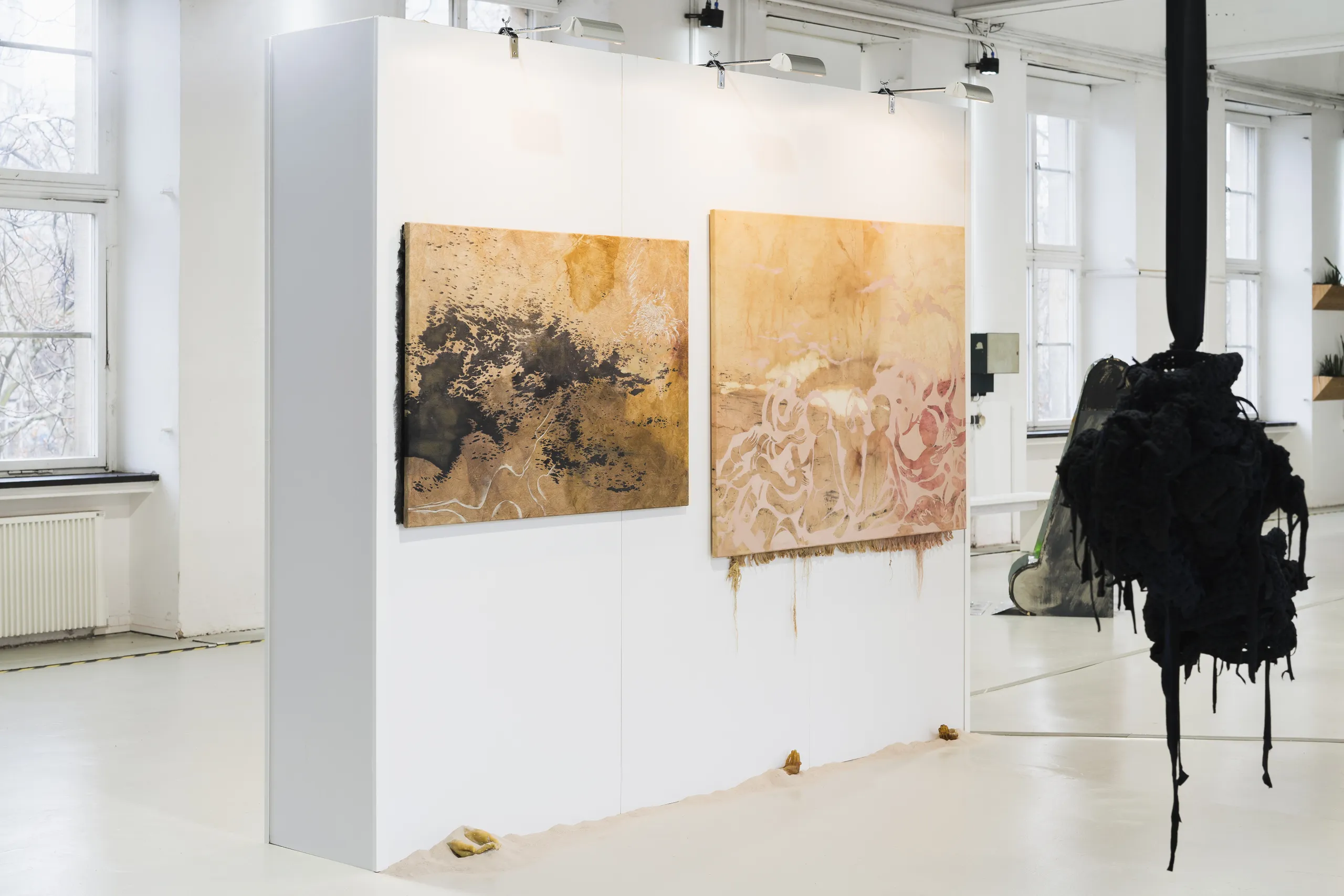 Das Bild zeigt die Ausstellungsansicht des ersten Rundgangs der Jüdischen Kunstschule. Zwei abstrakte Gemälde in Erdtönen hängen an einer weißen Stellwand in hellen Ausstellungsräumen mit großen Fenstern. Im Vordergrund hängt eine organisch wirkende Skulptur von der Decke.