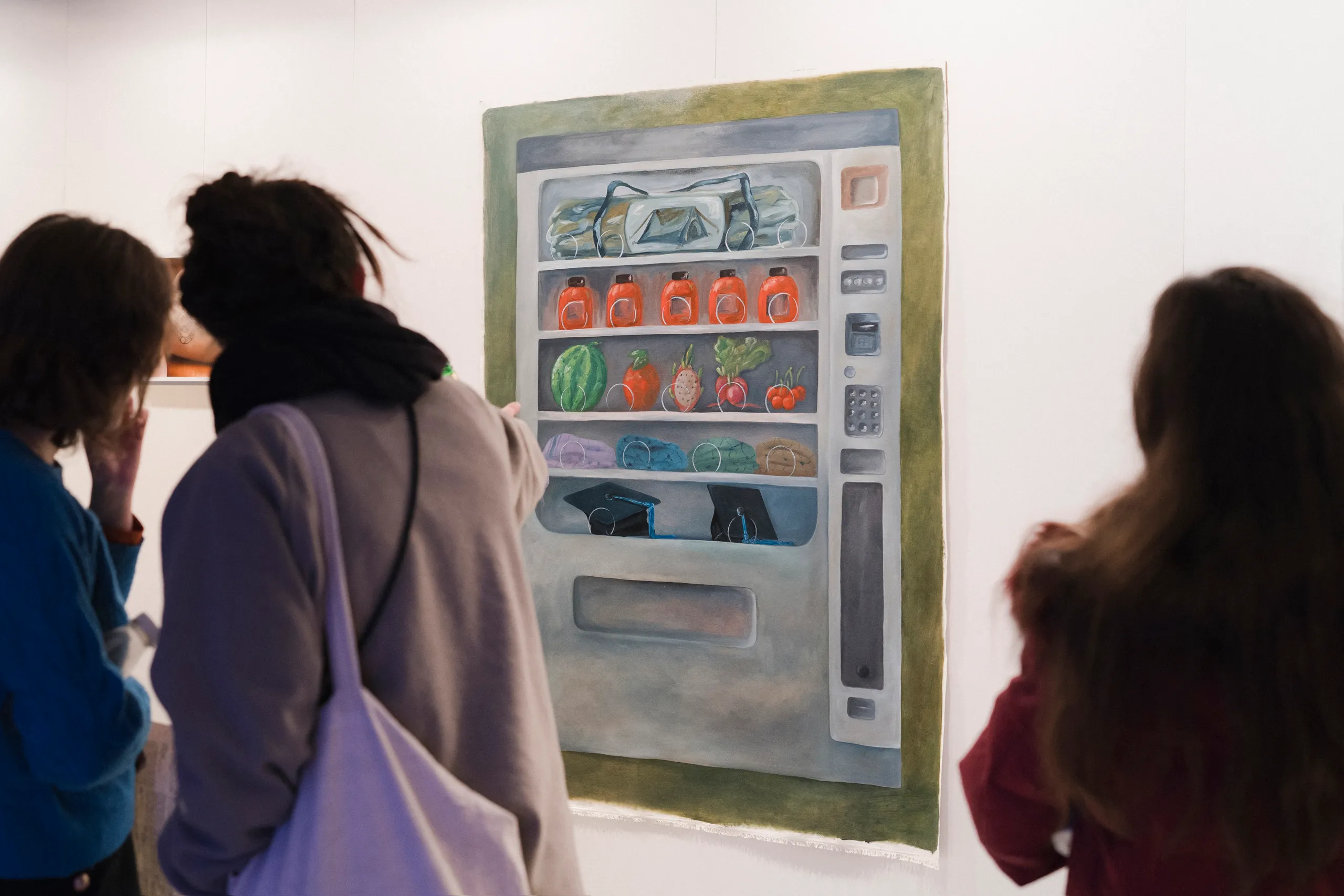 The picture shows the exhibition view of the first tour of the Jewish Art School. Three people are looking at a painting of a vending machine with water bottles, jam jars, fruit and vegetables in a gallery.